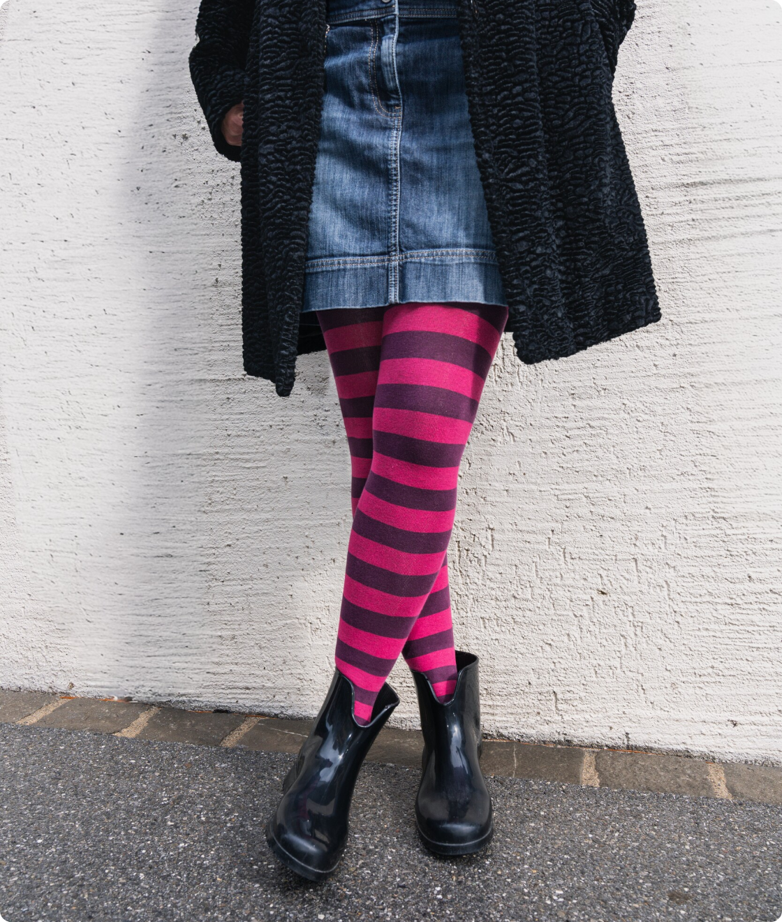 Person in dunklem Mantel, Jeansrock und schwarzen Stiefeletten trägt eine geringelte Strumpfhose in Pink und Dunkelviolett vor einer weissen Wand.