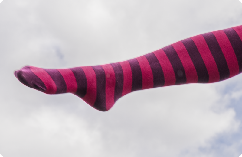 Person im Rock kickt ein Bein mit geringelter Strumpfhose in Pink und Dunkelviolett über einen Holzpfosten, das andere Bein steht auf einem zweiten Pfosten.