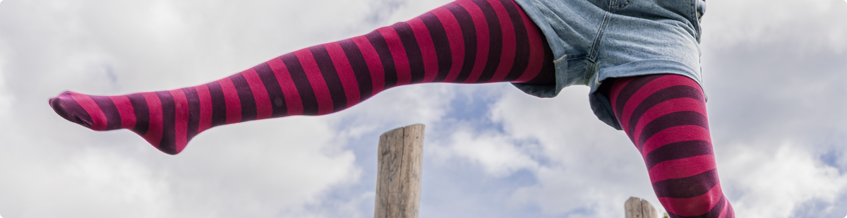 Person im Rock kickt ein Bein mit geringelter Strumpfhose in Pink und Dunkelviolett über einen Holzpfosten, das andere Bein steht auf einem zweiten Pfosten.
