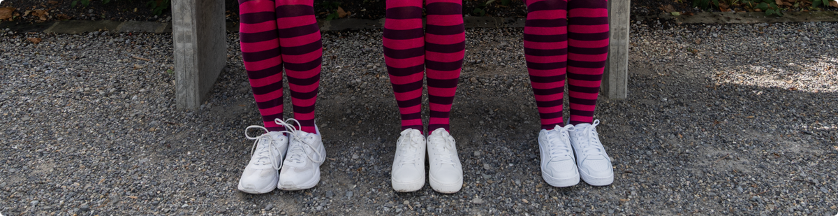 Drei Beinpaare in geringelten Strumpfhosen in Pink und Dunkelviolett und weissen Sneakers stehen nebeneinander auf einem Kiesweg.