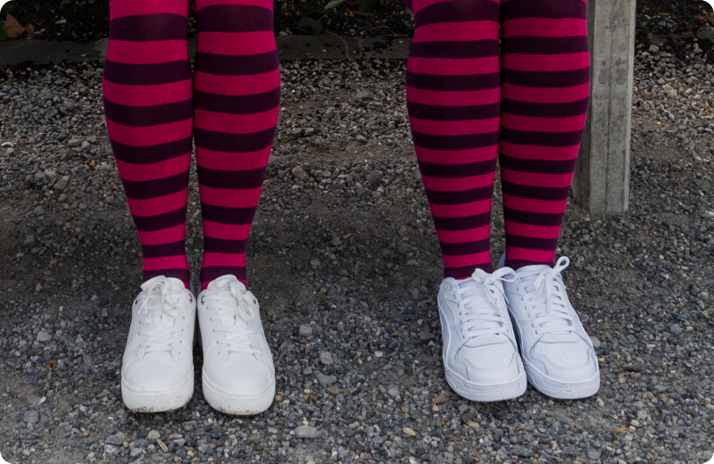 Person schaukelt auf einer Schaukel im Garten, trägt weisse Sneakers und eine geringelte Strumpfhose in Pink und Dunkelviolett.