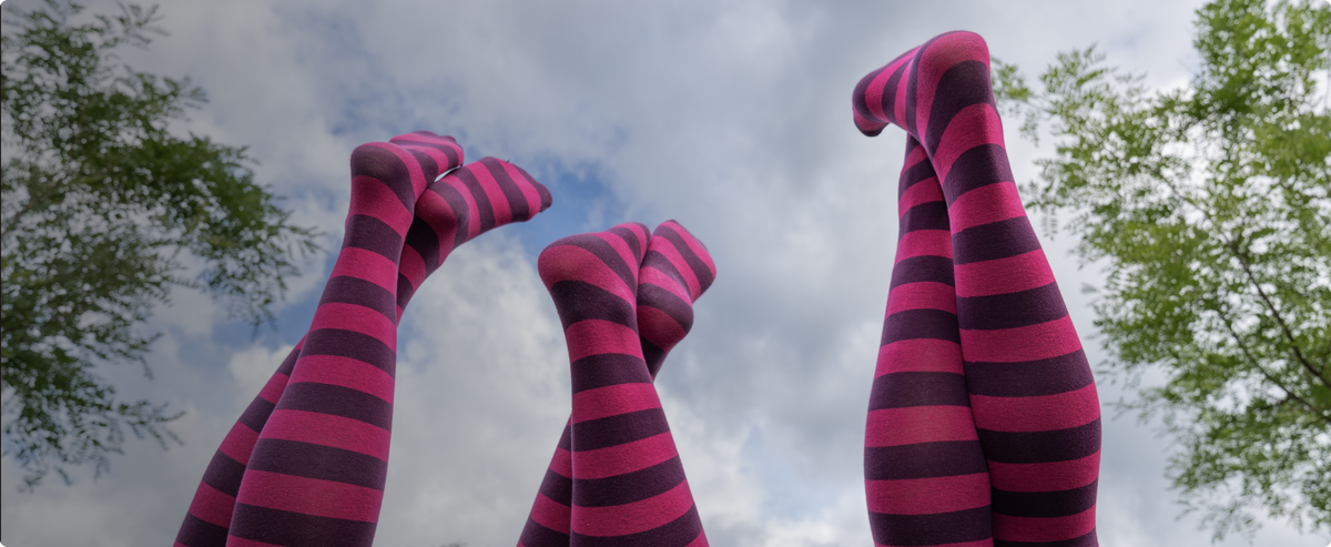 Mehrere Beine in geringelten Strumpfhosen in Pink und Dunkelviolett ragen vor bewölktem Himmel und Baumkronen in die Höhe.