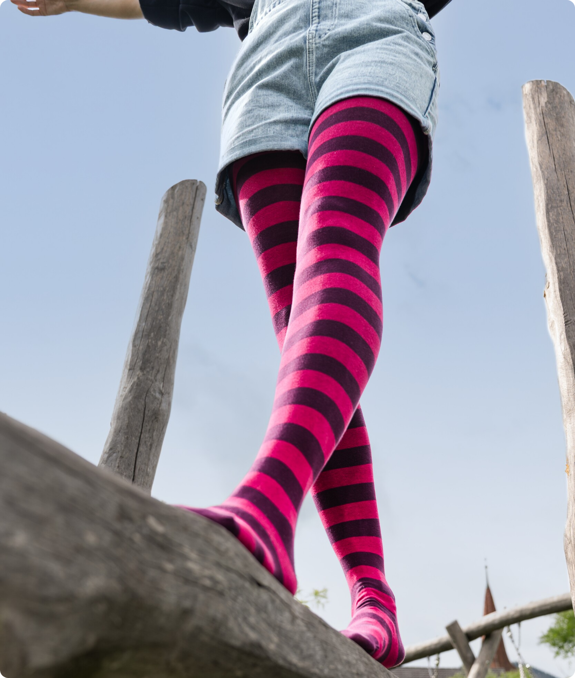 Person in Jeans-Shorts balanciert mit geringelter Strumpfhose in Pink und Dunkelviolett auf einem Baumstamm im Freien.