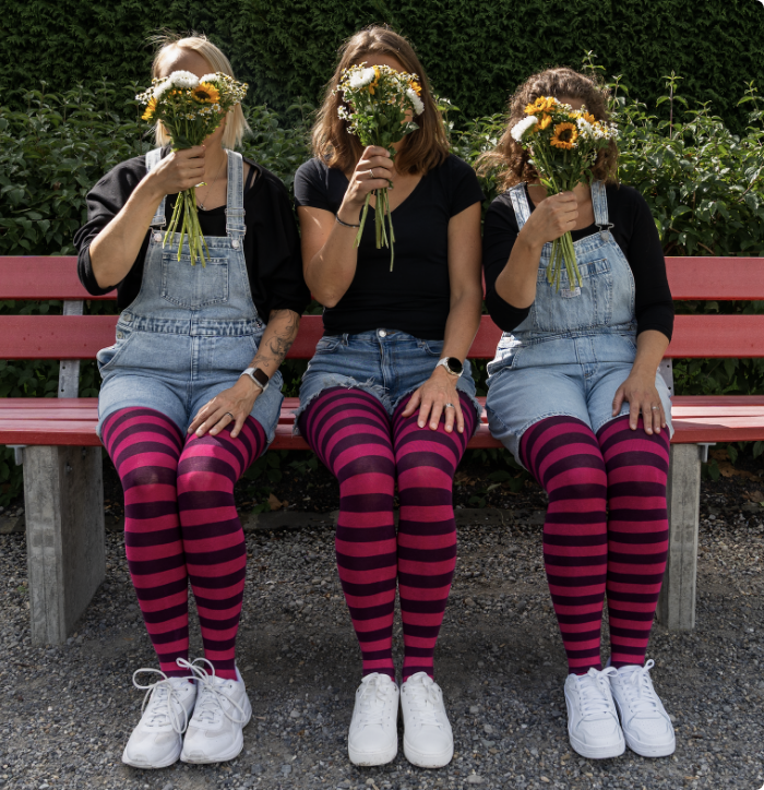 Drei Frauen in Jeans-Overalls stehen vor einer grünen Hecke und halten sich Blumensträusse vors Gesicht, alle tragen geringelte Strumpfhosen in Pink und Dunkelviolett zu weissen Sneakers.
