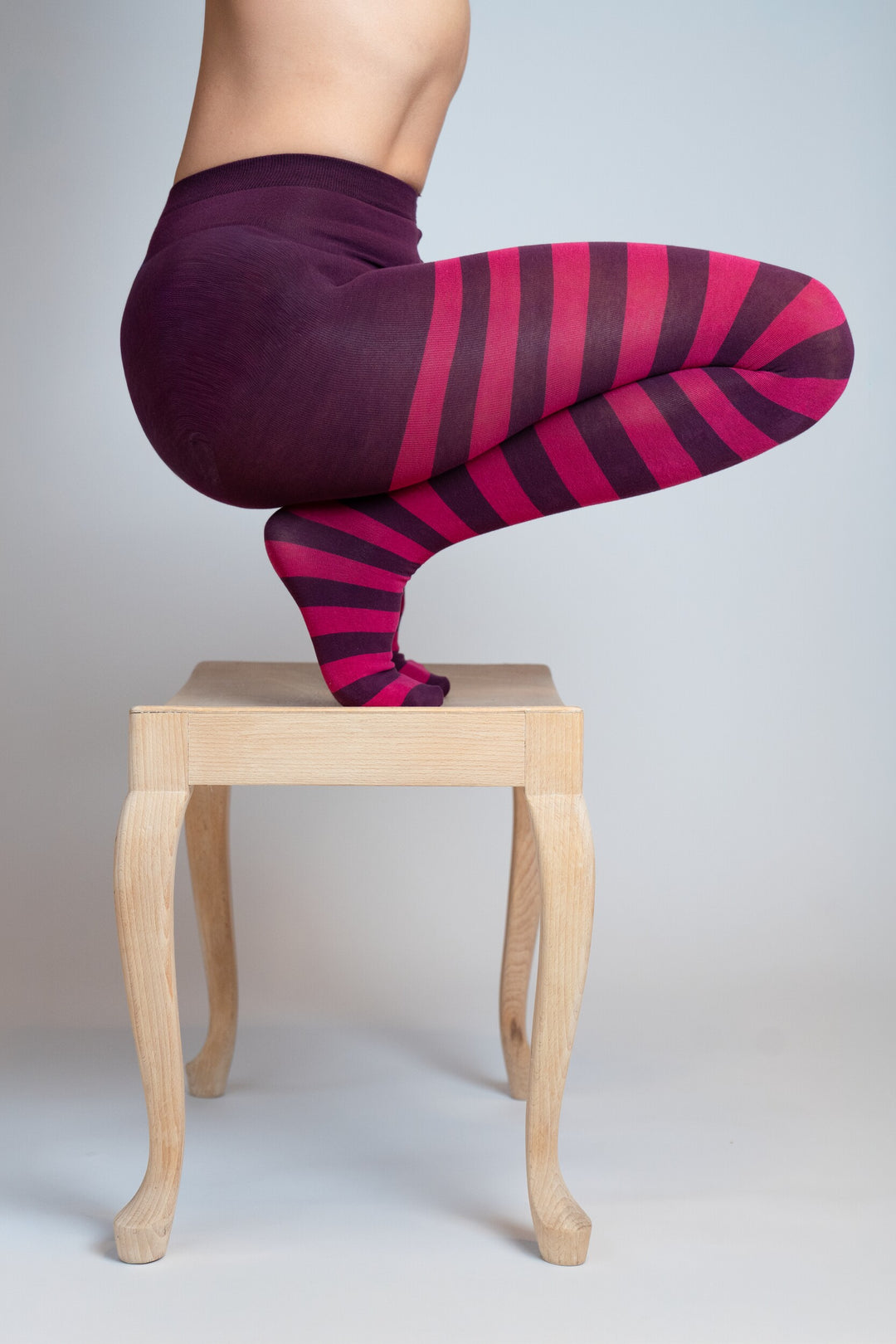Strumpfhose mit dunkelviolettem Bund und Ringelmuster in Pink und Dunkelviolett, in hockender Pose auf einem kleinen Holzhocker, auf hellem Hintergrund.