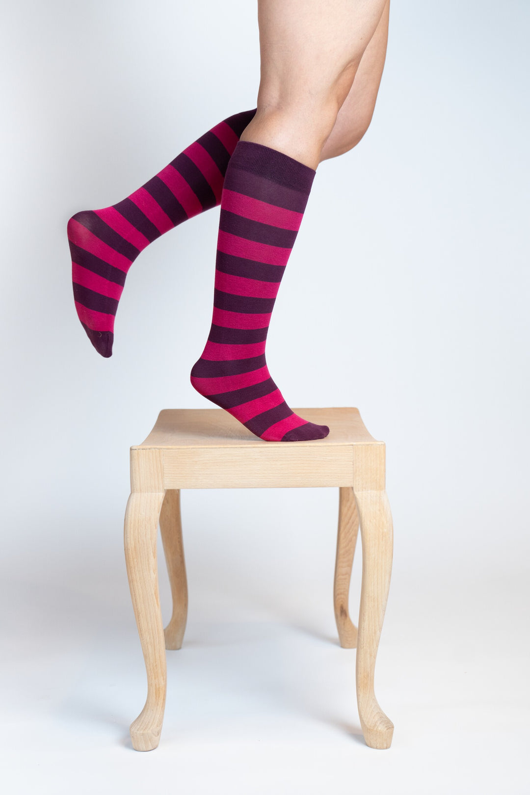 Zwei Beine mit geringelten Kniestrümpfen in Pink und Dunkelviolett, auf Zehenspitzen stehend auf einem kleinen Holzhocker, vor hellem Hintergrund.