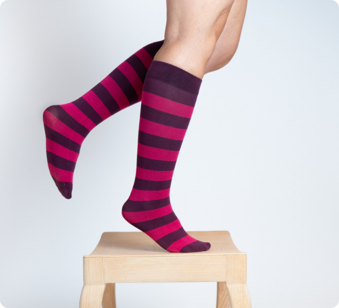 Zwei Beine mit geringelten Kniestrümpfen in Pink und Dunkelviolett, auf Zehenspitzen stehend auf einem kleinen Holzhocker, vor hellem Hintergrund.