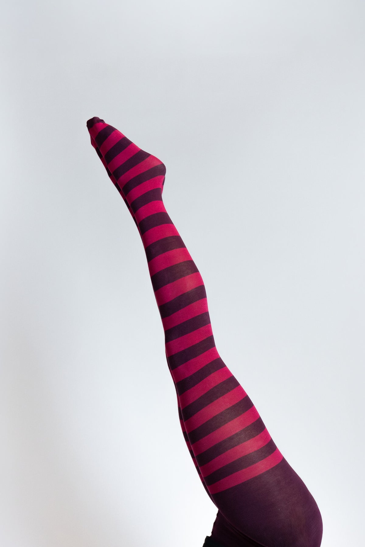 Hoch gestrecktes Bein mit geringelter Strumpfhose in Pink und Dunkelviolett und gestreckter Fussspitze, auf hellem Hintergrund.