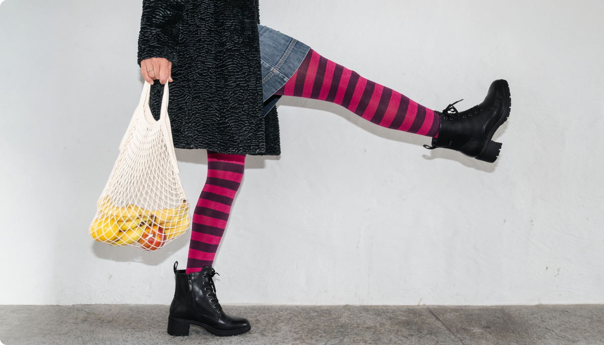 Frau in dunklem Mantel hält einen Einkaufsbeutel mit Früchten und kickt ein Bein mit geringelter Strumpfhose in Pink und Dunkelviolett nach hinten.