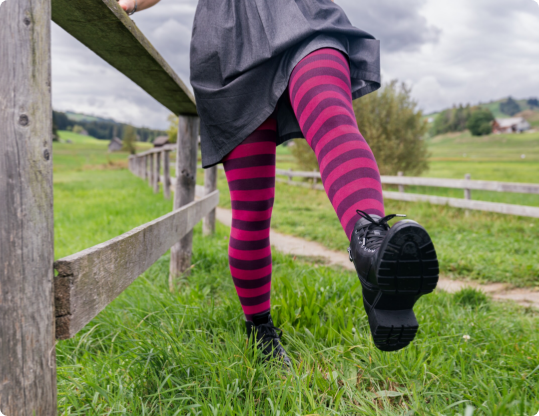 Person im dunklen Rock klettert über einen Holzzaun, trägt schwarze Stiefel und eine geringelte Strumpfhose in Pink und Dunkelviolett.